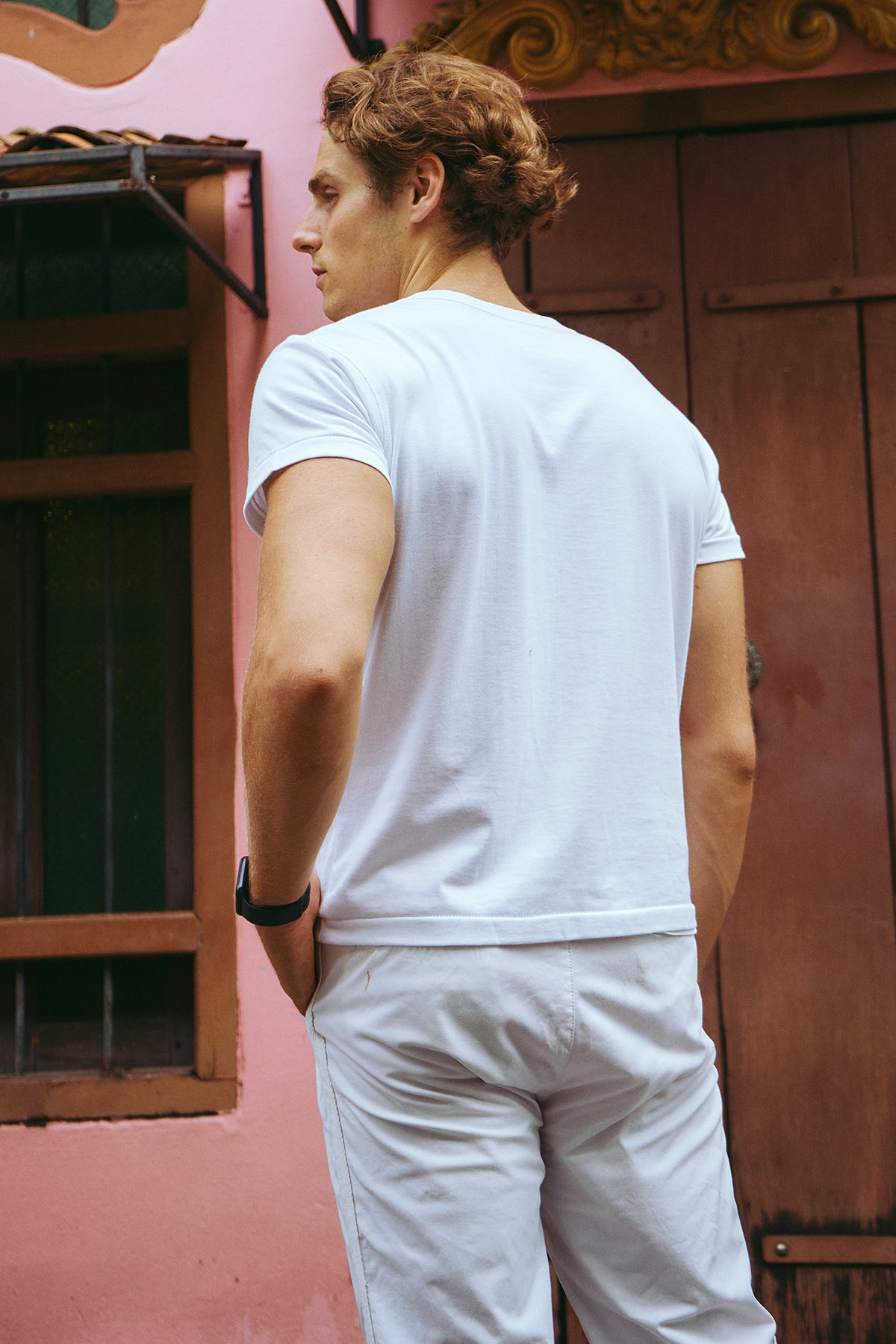 A man wearing a white short-sleeve crewneck t-shirt and white pants, shown from behind, standing in front of a pink wall and dark wooden door.