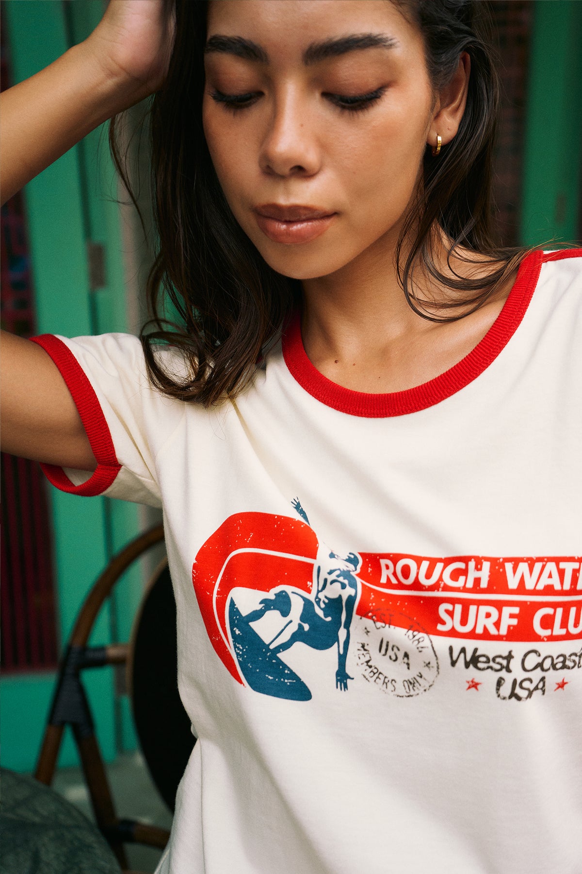 A woman wearing a white ringer t-shirt with red trim and a graphic print that reads 'ROUGH WATER SURF CLUB West Coast USA'.