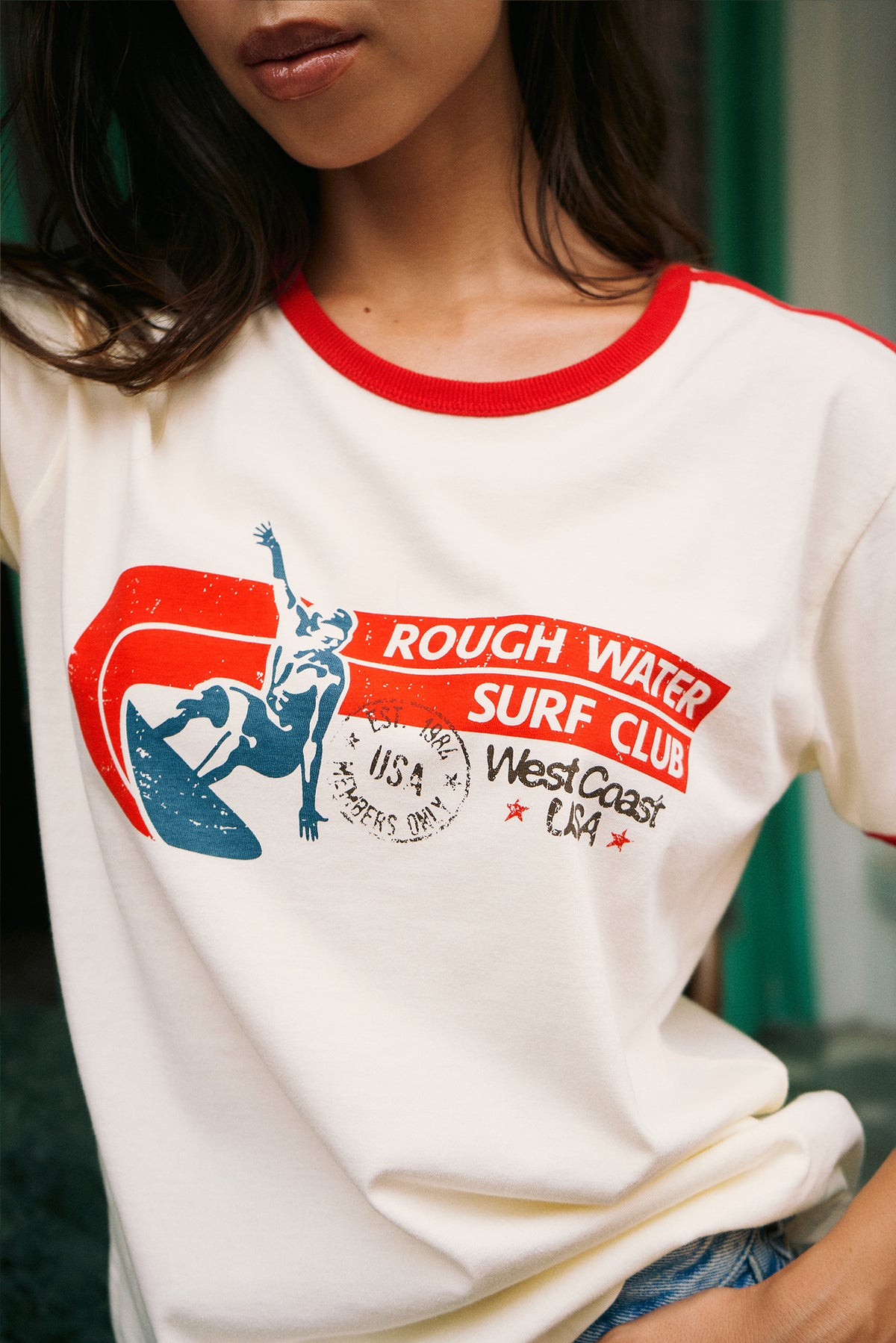A close-up of a person wearing a cream ringer t-shirt with red trim and a red, white, and blue graphic that reads 'ROUGH WATER SURF CLUB' and 'West Coast USA'.