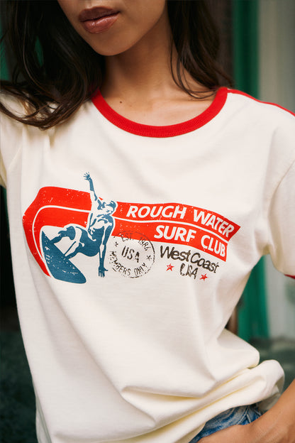 A close-up of a person wearing a cream ringer t-shirt with red trim and a red, white, and blue graphic that reads 'ROUGH WATER SURF CLUB' and 'West Coast USA'.