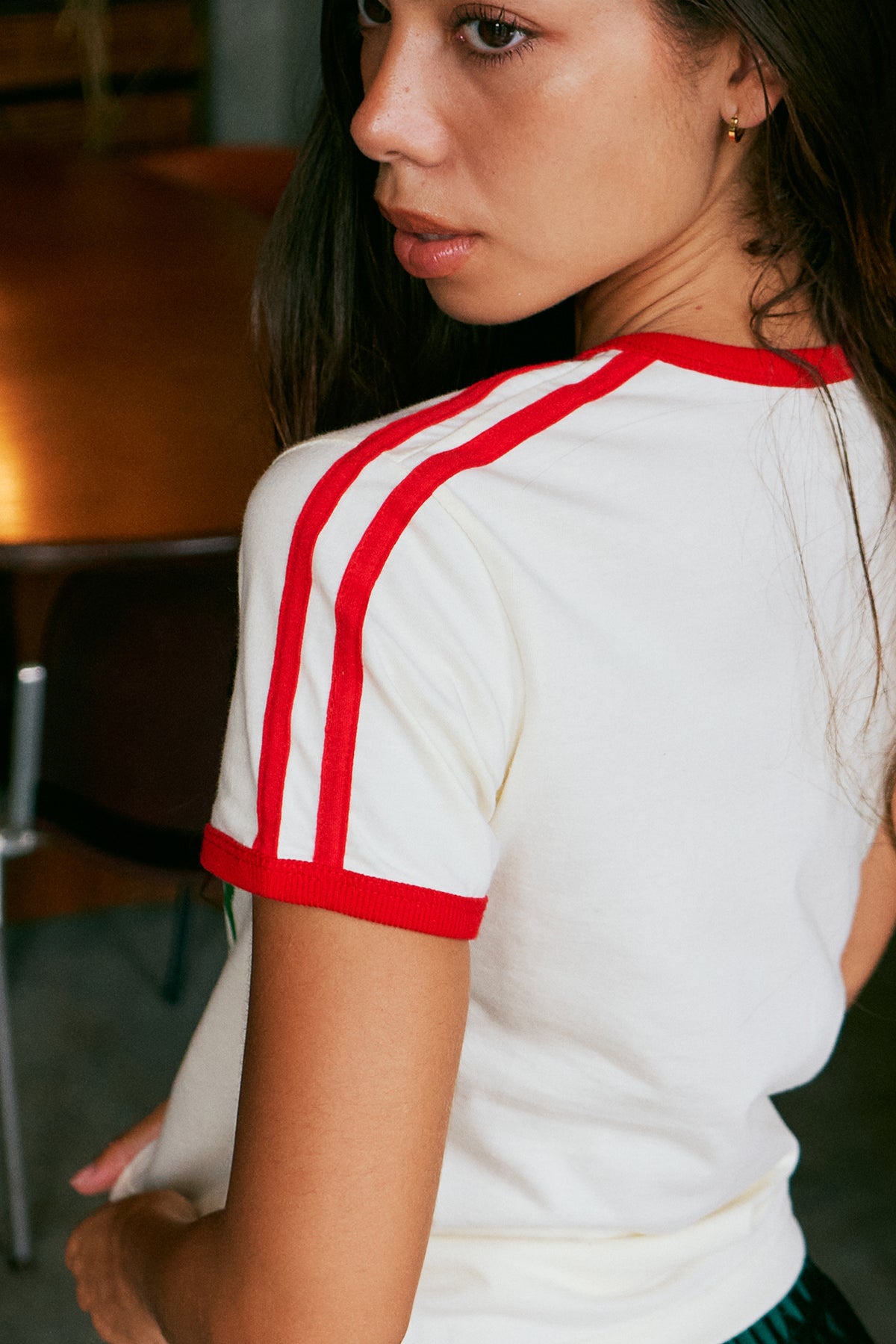 A close-up lifestyle shot of a woman wearing a white short-sleeve t-shirt with red contrast trim on the collar and sleeve cuffs, featuring double red stripes down the right shoulder.