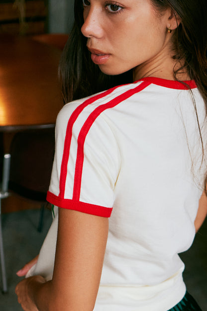 A close-up lifestyle shot of a woman wearing a white short-sleeve t-shirt with red contrast trim on the collar and sleeve cuffs, featuring double red stripes down the right shoulder.