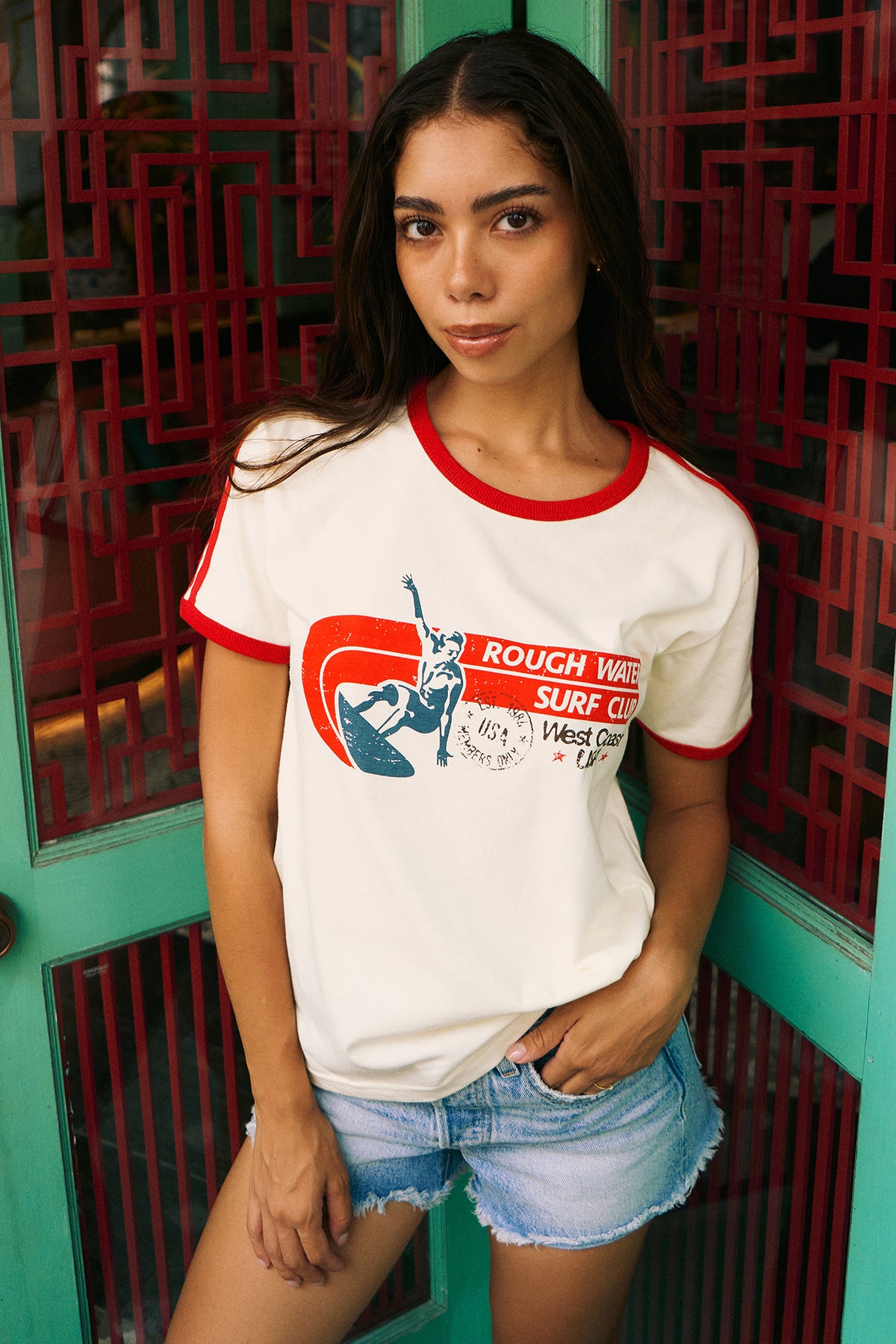 A woman wearing a white ringer t-shirt with red trim and a retro graphic that reads 'ROUGH WAVE SURF CLUB West Coast', paired with denim shorts, standing in front of a teal and red geometric door.