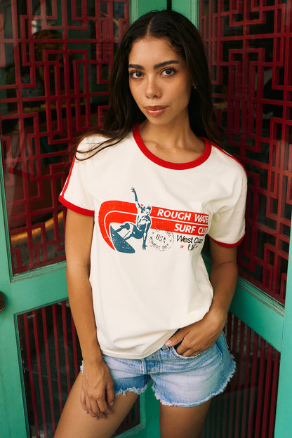 A woman wearing a white ringer t-shirt with red trim and a retro graphic that reads 'ROUGH WAVE SURF CLUB West Coast', paired with denim shorts, standing in front of a teal and red geometric door.