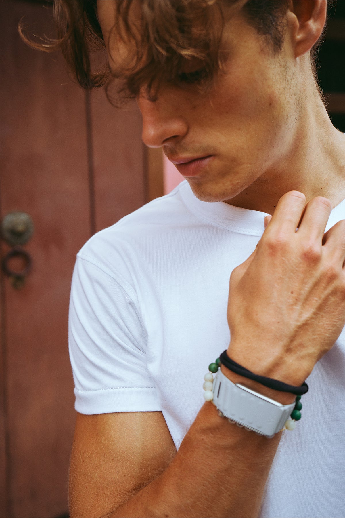 A close-up lifestyle shot of a man wearing a white crew-neck t-shirt and a white smartwatch with a black band and green beads on his wrist, set against a rustic reddish-brown wooden door background.