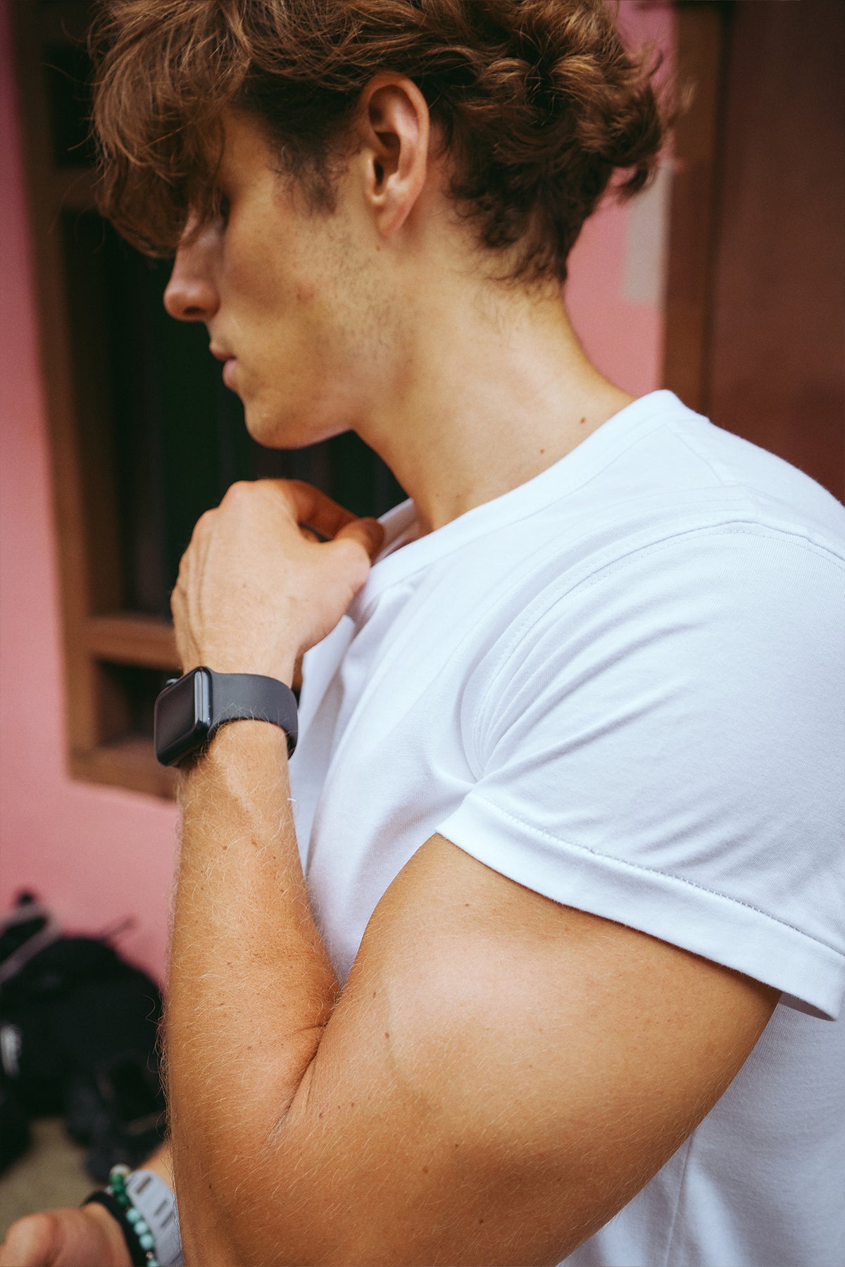 A close-up side profile of a man wearing a white short-sleeve crewneck t-shirt and a black smartwatch, standing against a pink wall.