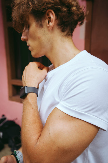 A close-up side profile of a man wearing a white short-sleeve crewneck t-shirt and a black smartwatch, standing against a pink wall.