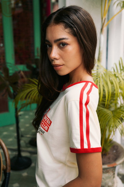 A woman wearing a white ringer t-shirt with red trim and red stripes on the sleeves, posed in an outdoor patio setting with green plants in the background.