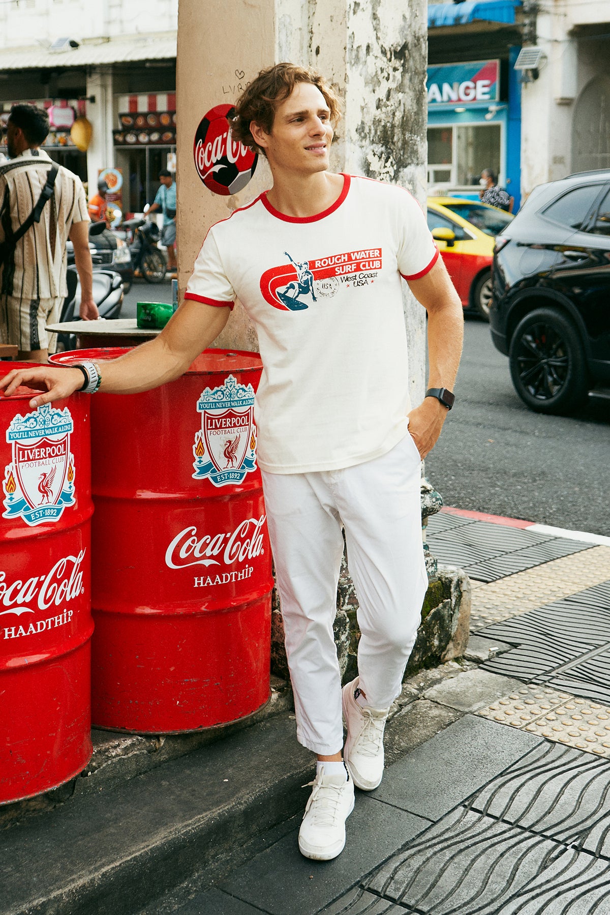 A man wearing a white ringer t-shirt with red trim and a red, white, and blue 'Rough Water Surf Club' graphic on the chest, standing on a city sidewalk next to a street and bakery storefront.