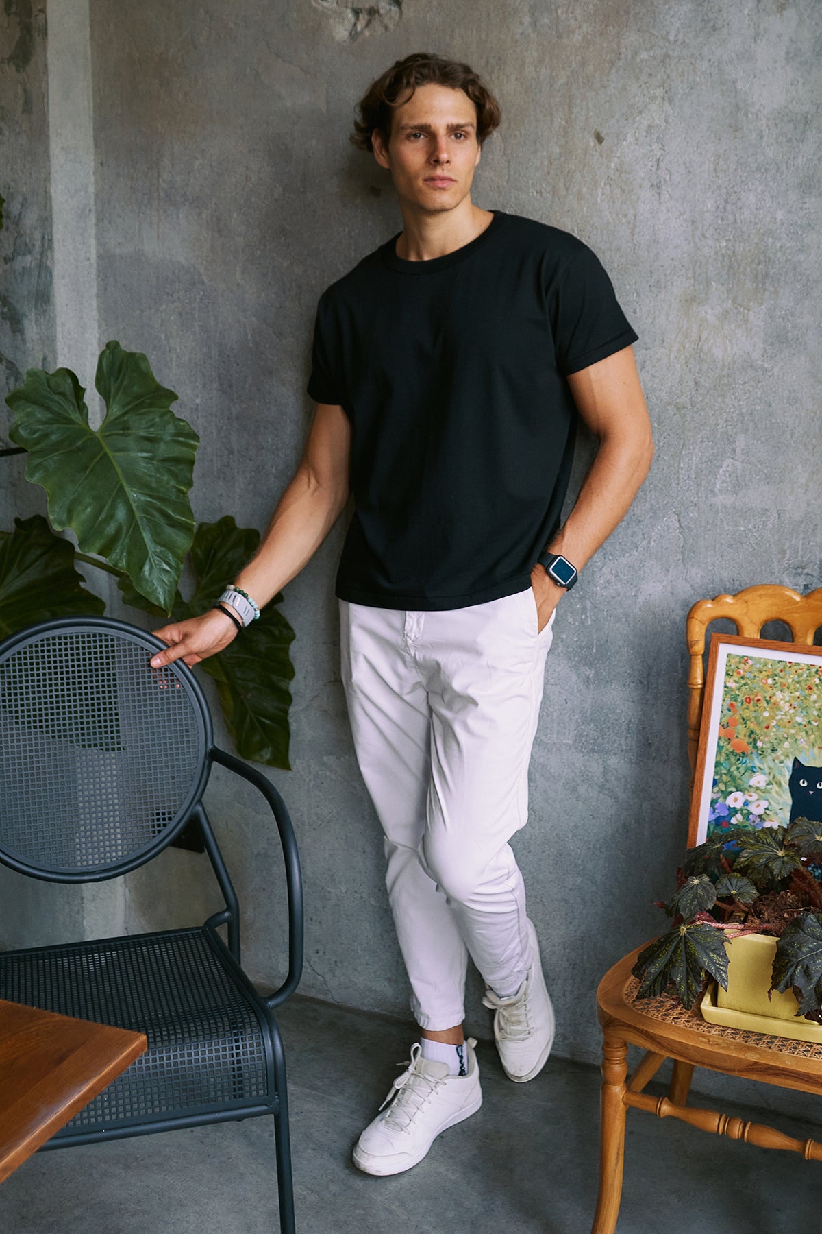 A man wearing a black crewneck t-shirt, white pants, and white sneakers stands in a room with a textured gray wall, leaning against a black metal chair next to a wooden table with plants.