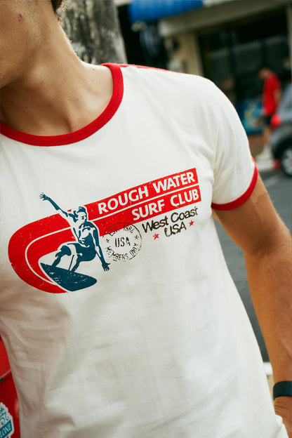 A man wearing a white ringer t-shirt with red trim and a red, white, and blue 'Rough Water Surf Club' graphic on the chest, standing on a city sidewalk next to a street and bakery storefront.