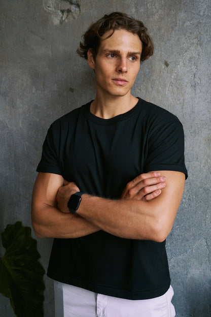 A man with wavy brown hair wearing a black short-sleeve crewneck t-shirt and white pants, standing with arms crossed against a textured gray concrete wall.