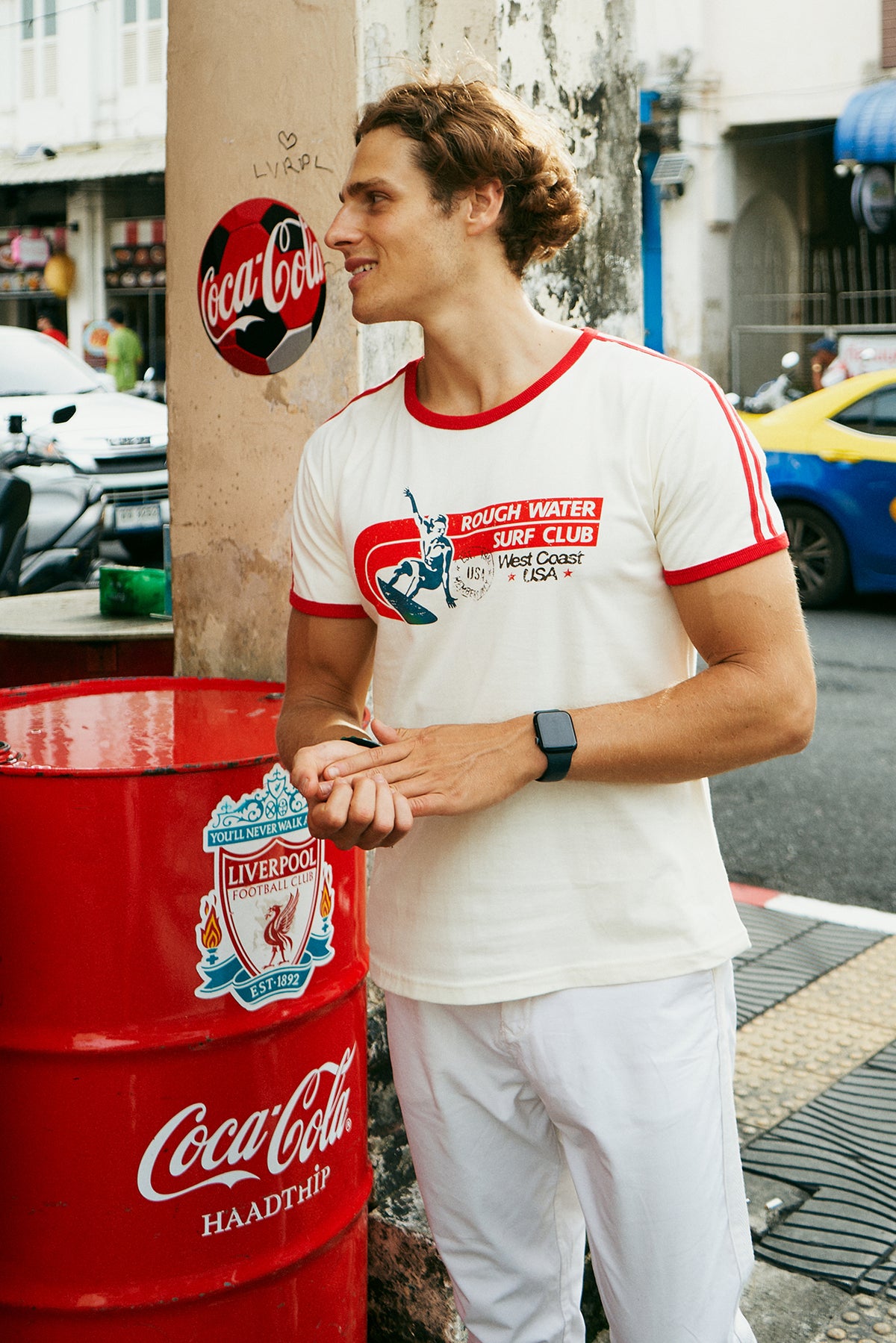 A man wearing a white ringer t-shirt with red trim and a red, white, and blue 'Rough Water Surf Club' graphic on the chest, standing on a city sidewalk next to a street and bakery storefront.