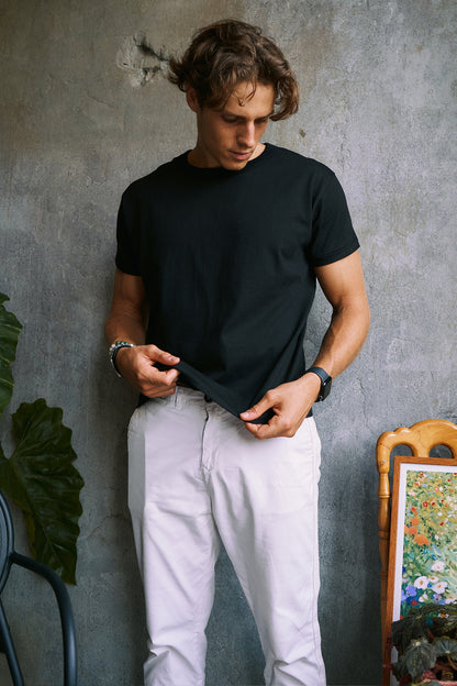 A man wearing a black short-sleeve crewneck t-shirt and off-white trousers, standing against a textured gray concrete wall.