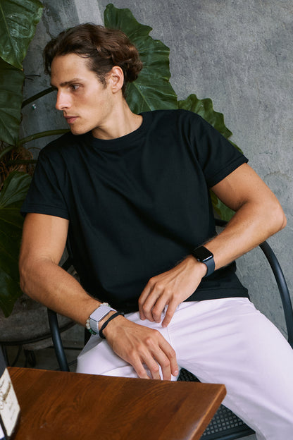 A man wearing a black short-sleeve crewneck t-shirt and white pants, sitting in a chair in front of a gray concrete wall with green leaves.
