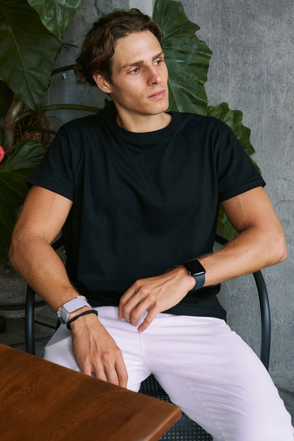 A man wearing a black short-sleeve crewneck t-shirt and white pants, sitting in a chair in front of a gray concrete wall with green leaves.