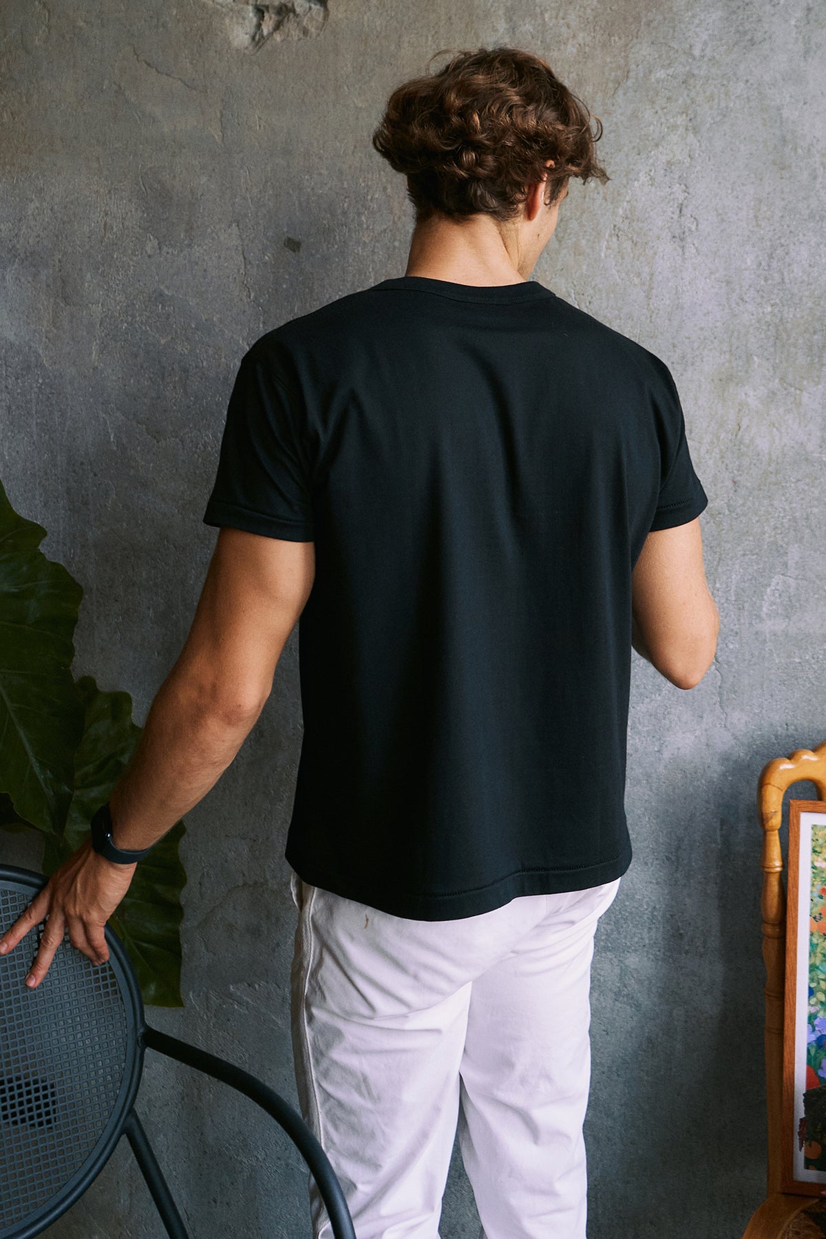 A man viewed from behind wearing a plain black short-sleeve t-shirt and white pants, standing against a textured gray wall.
