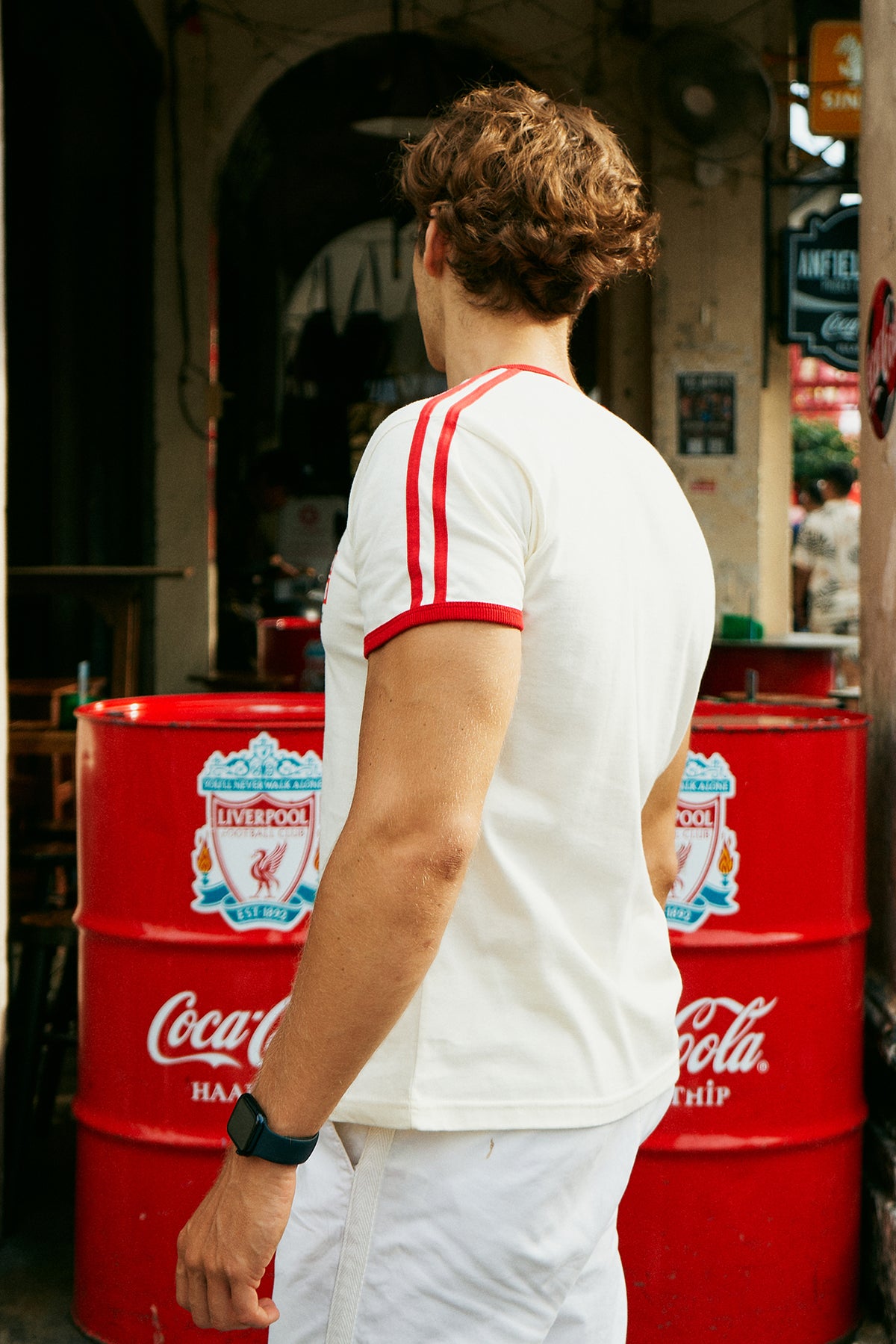 A man wearing a white ringer t-shirt with red trim and a red, white, and blue 'Rough Water Surf Club' graphic on the chest, standing on a city sidewalk next to a street and bakery storefront.