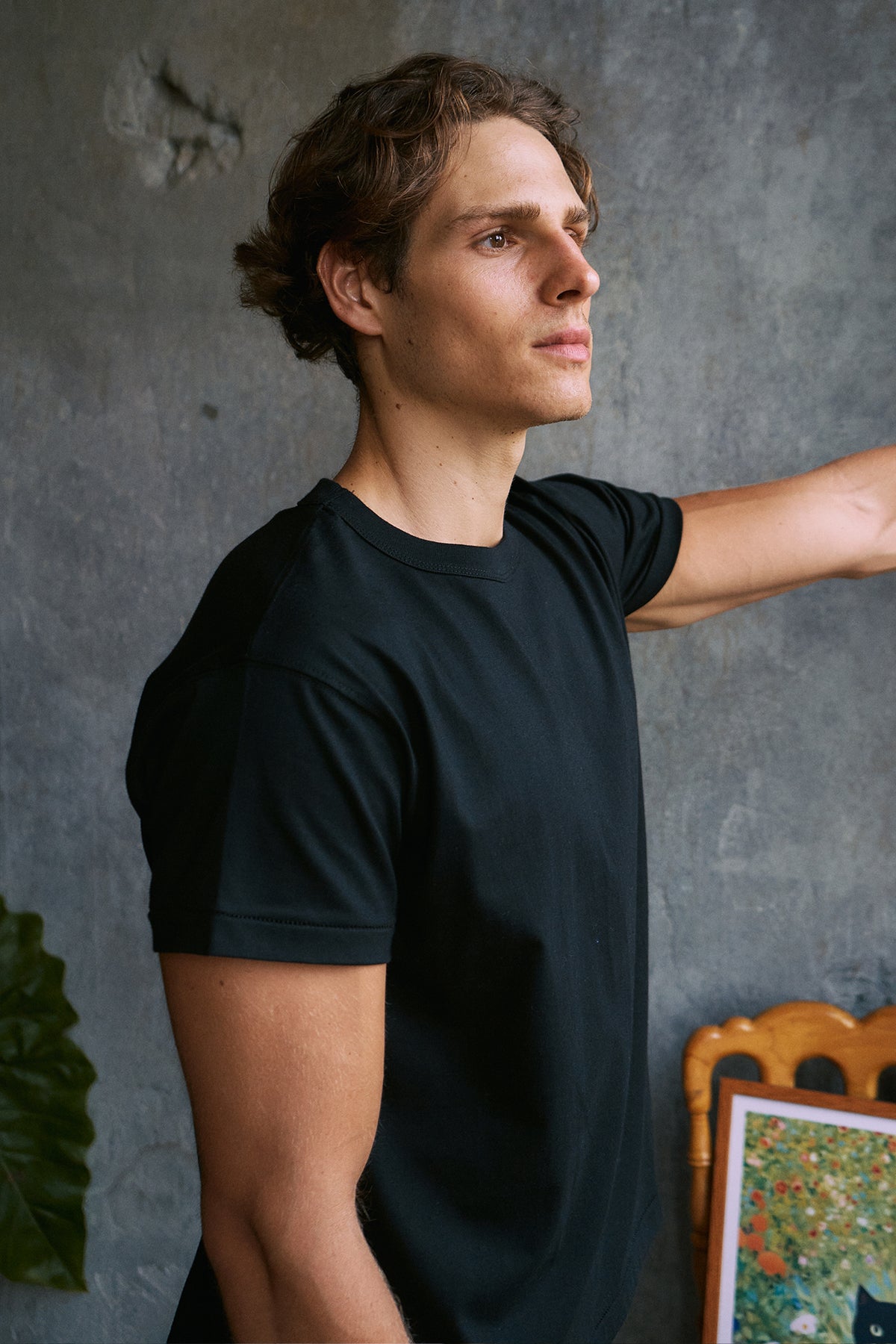 A man wearing a plain black short-sleeve crewneck t-shirt, standing against a textured gray wall.
