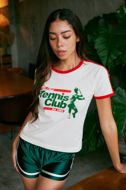 A woman wearing a white short-sleeve graphic T-shirt with red trim and a green 'Tennis Club' print, paired with dark green athletic shorts, posed in a cafe setting with large green leaves in the background.