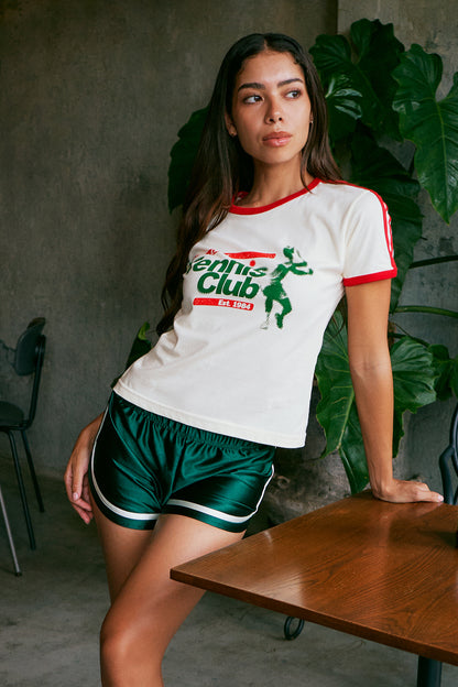 A woman leans against a wooden table in a cafe setting, wearing a white ringer t-shirt with red trim and a green 'Tennis Club' graphic, paired with shiny dark green athletic shorts.