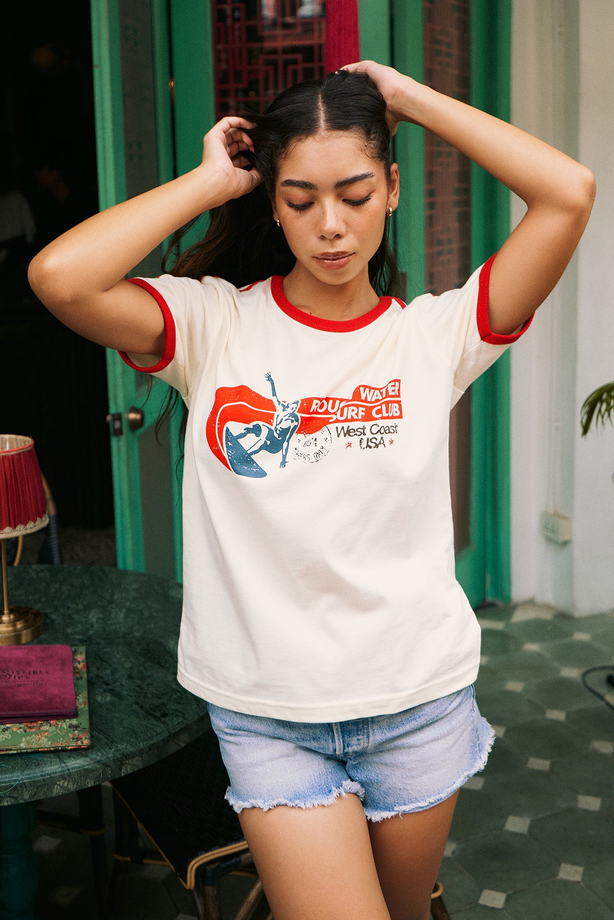 A woman wearing a white ringer t-shirt with red trim and a red, white, and blue 'Rough Water Surf Club' graphic on the chest, standing on a city sidewalk next to a street and bakery storefront.