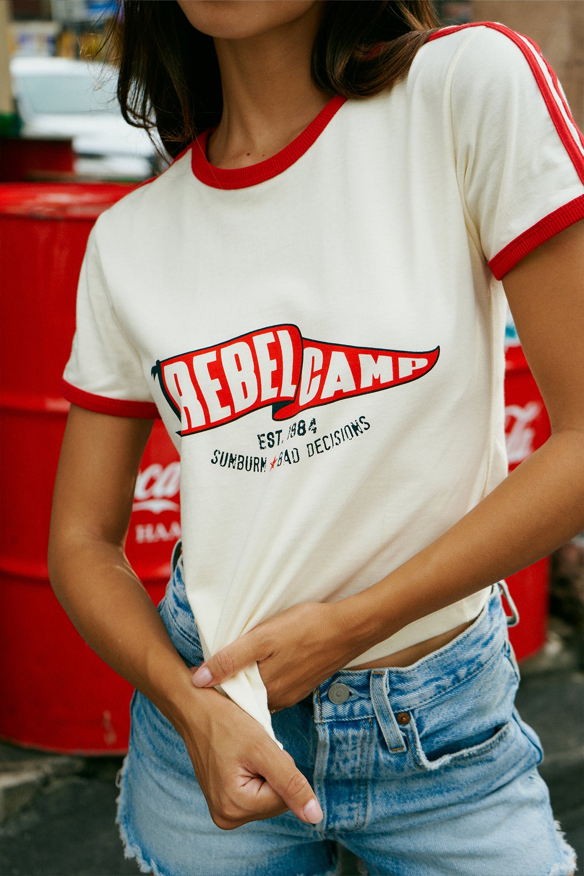 A woman wearing a cream ringer t-shirt with red trim and a red, black, and blue 'Rebel Camp' graphic on the front, paired with light-wash blue jeans.