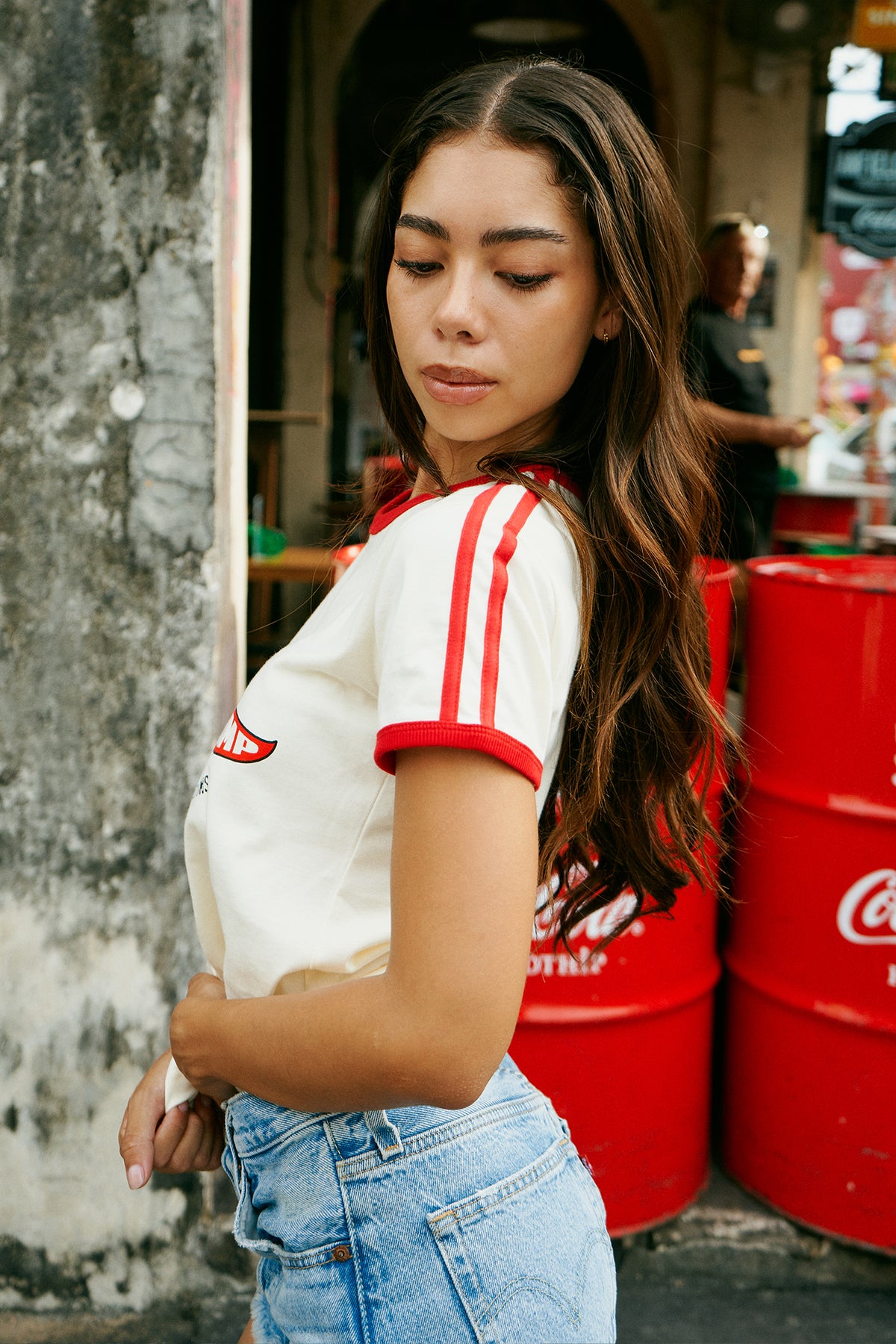 A woman wearing a cream ringer t-shirt with red trim and a 'Rebel Camp' graphic, paired with denim shorts, leaning against a weathered wall next to a red Coca-Cola container