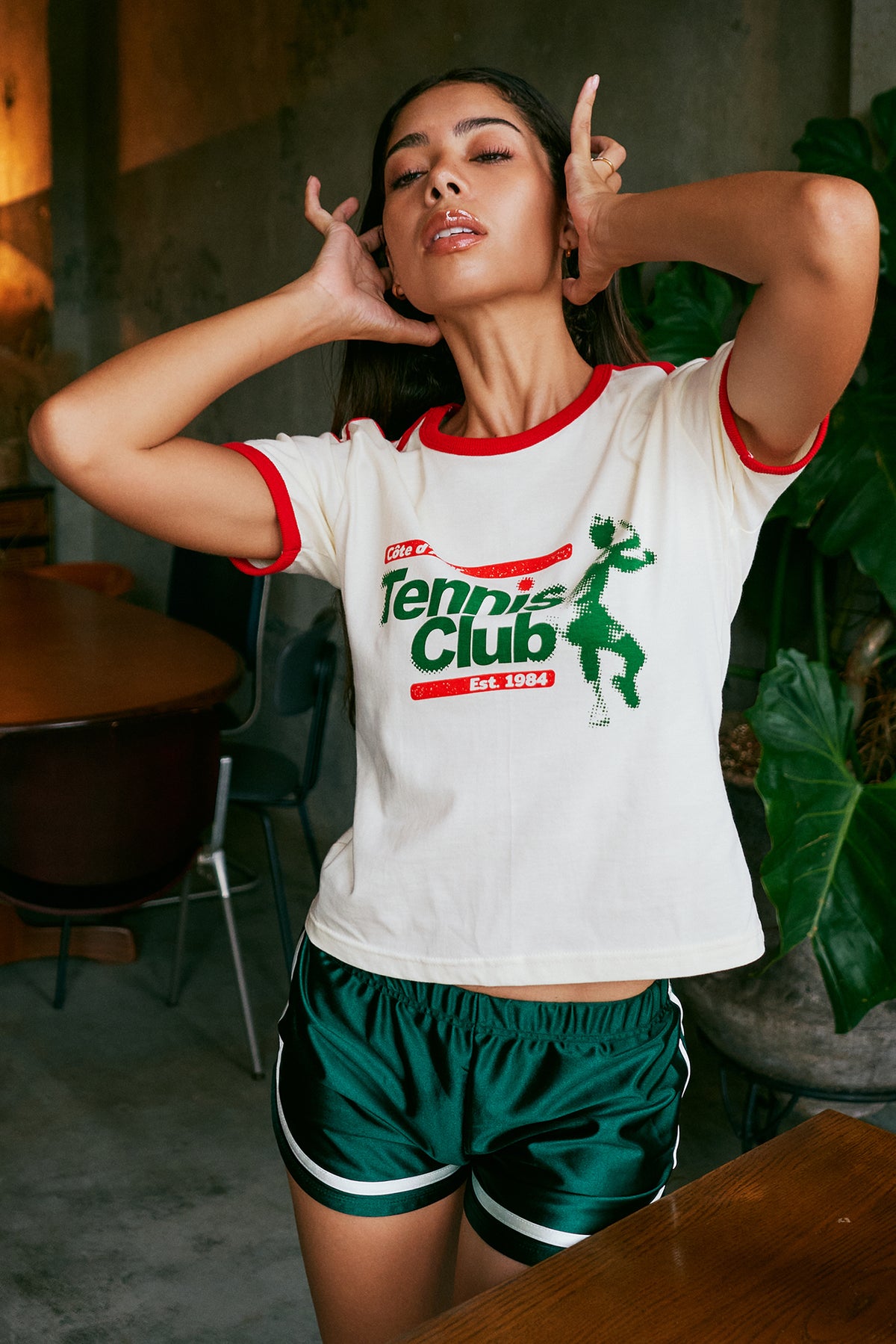 A woman posing in a white ringer t-shirt with red and green trim and a 'Tennis Club' graphic, paired with green athletic shorts in a cafe setting.