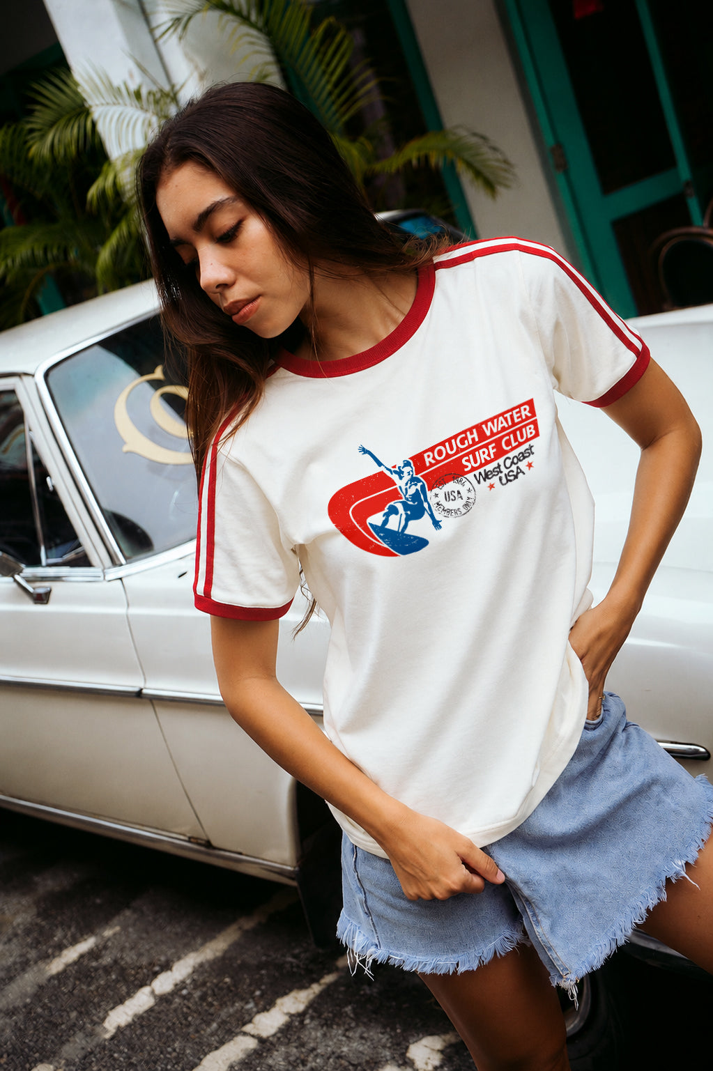 A woman wearing a white short-sleeve ringer T-shirt with red trim and a retro graphic on the chest, paired with denim shorts, standing next to a white vintage car outdoors.