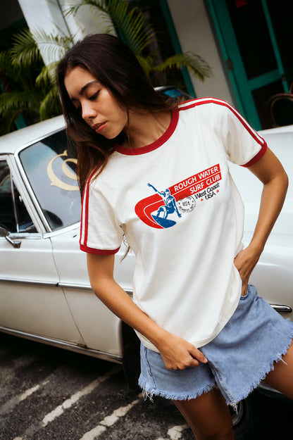 A woman wearing a white short-sleeve ringer T-shirt with red trim and a retro graphic on the chest, paired with denim shorts, standing next to a white vintage car outdoors.