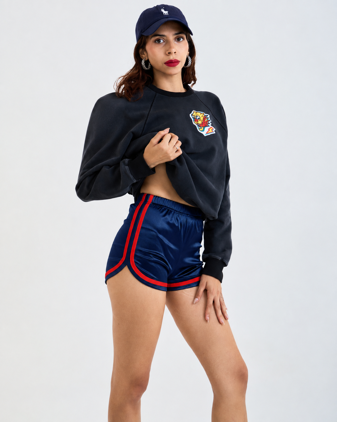 A model wearing a navy blue cropped sweatshirt with a colorful tiger graphic on the chest, matching navy athletic shorts with red and blue side stripes, and a navy baseball cap, posed against a light gray studio background.