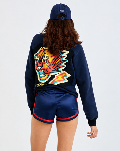 A model shown from the back wearing a navy blue pullover sweatshirt with a large colorful tiger graphic on the back, matching navy shorts with red trim, and a navy cap with white Polo branding.