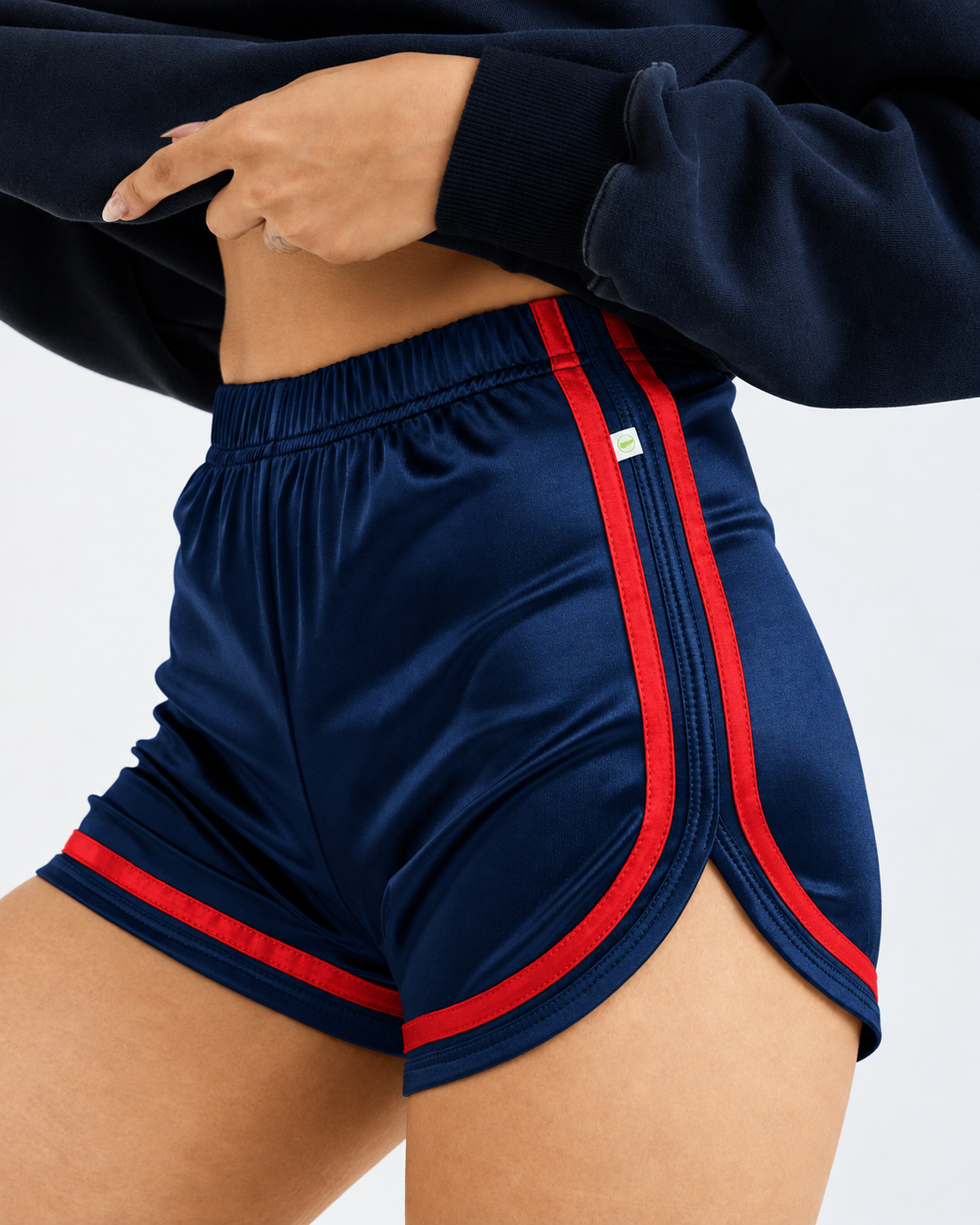 A close-up of a model wearing navy blue athletic shorts with red side and hem stripes, paired with a dark navy sweatshirt against a light gray studio background.