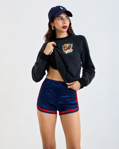 A woman modeling a black cropped sweatshirt with a colorful eagle patch on the chest, paired with navy blue athletic shorts featuring red side stripes, a navy cap, and hoop earrings, against a light gray studio background.