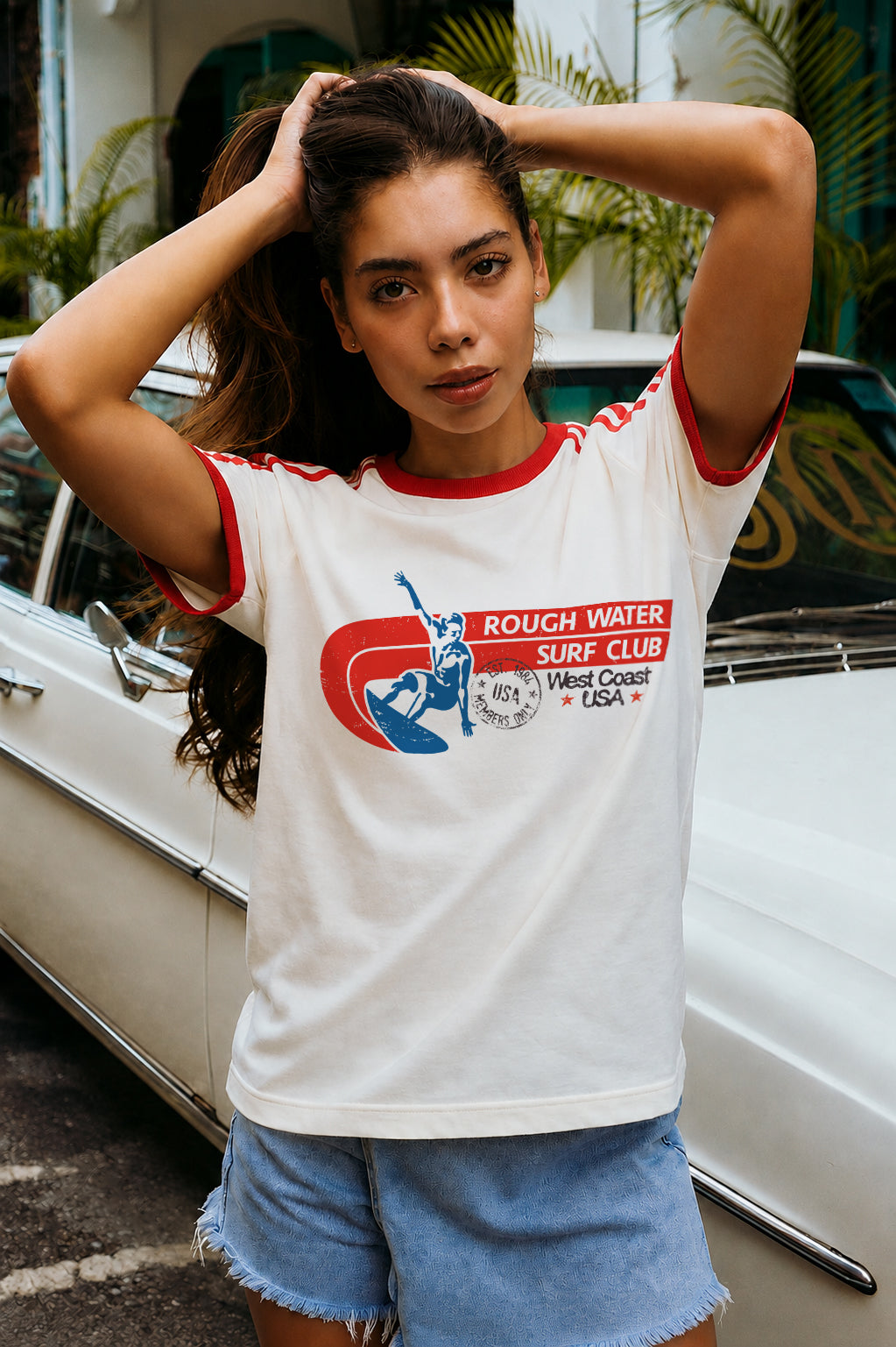A woman wearing a white ringer t-shirt with red trim and a retro graphic that reads 'ROUGH WATER SURF CLUB West Coast USA', standing in front of a white vintage car.