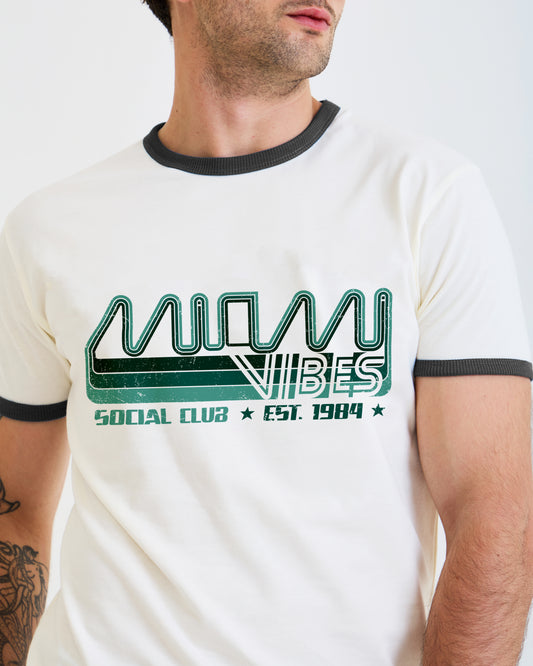 A man wearing a cream-colored ringer t-shirt with a green and black graphic on the chest that reads 'MUMBLE VIBES SOCIAL CLUB EST. 1984'.
