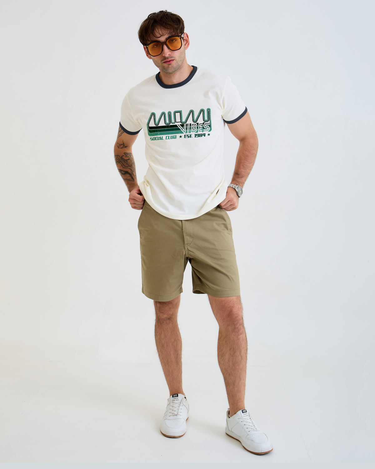 A man modeling a white ringer t-shirt with green and navy lettering, paired with khaki shorts and white sneakers, standing against a plain white studio background.