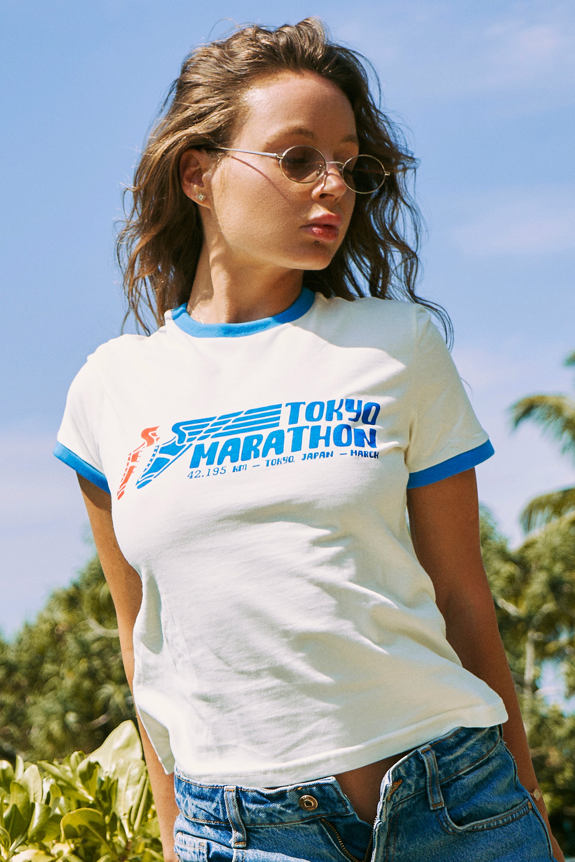 A woman wearing a white ringer T-shirt with blue trim and a 'Tokyo Marathon' graphic on the front, paired with blue jeans, photographed outdoors against a blue sky and green foliage background.