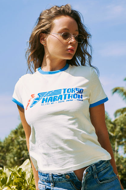 A woman wearing a white ringer T-shirt with blue trim and a 'Tokyo Marathon' graphic on the front, paired with blue jeans, photographed outdoors against a blue sky and green foliage background.
