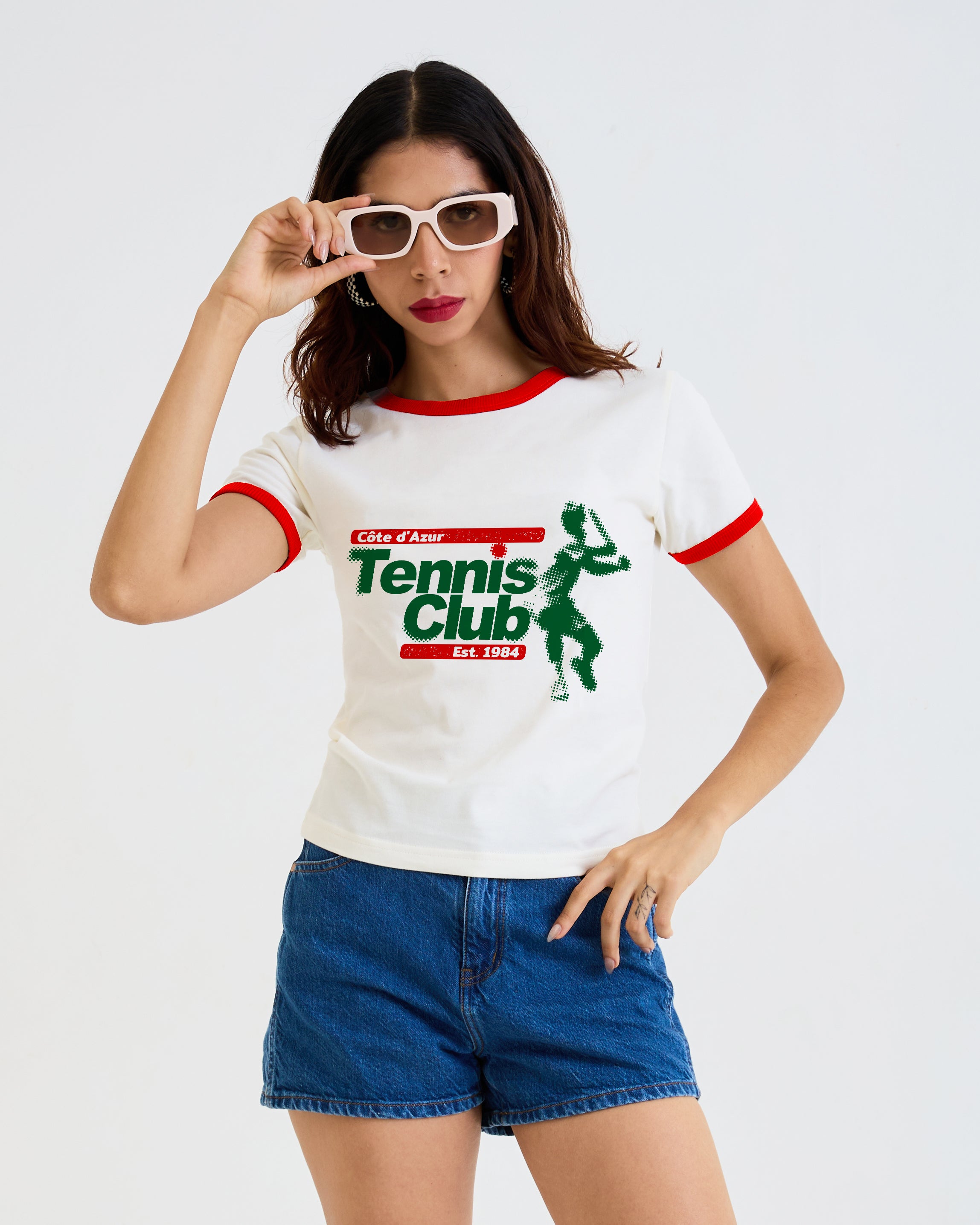 A woman modeling a white ringer T-shirt with green and red trim and a 'Tennis Club' graphic, paired with blue denim shorts against a light gray studio background.