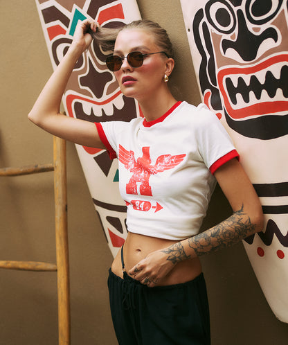 Woman wearing a white crop top with a red eagle design in front of tribal-style wall art.