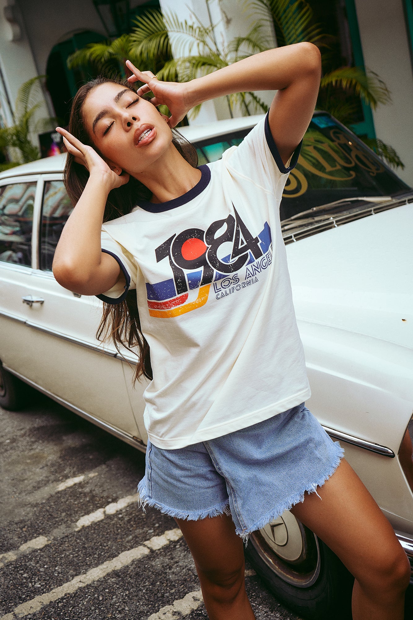 A woman posing in front of a vintage white car, wearing a white ringer t-shirt with a colorful retro '1984 Los Angeles' graphic and light blue denim shorts.