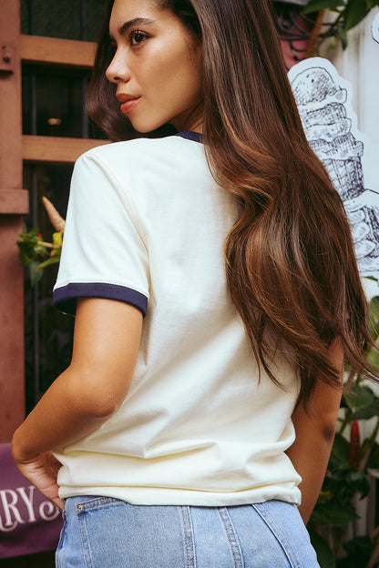 A woman with long brown hair is shown from the back wearing a cream-colored ringer t-shirt with dark navy blue trim on the collar and sleeve cuffs, paired with light blue jeans.