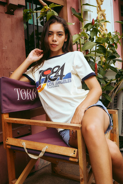 A woman sits in a wooden chair with a purple cushion, wearing a white graphic t-shirt with navy trim and a colorful Los Angeles graphic. The background features a rustic pinkish-brown wall and green plants.