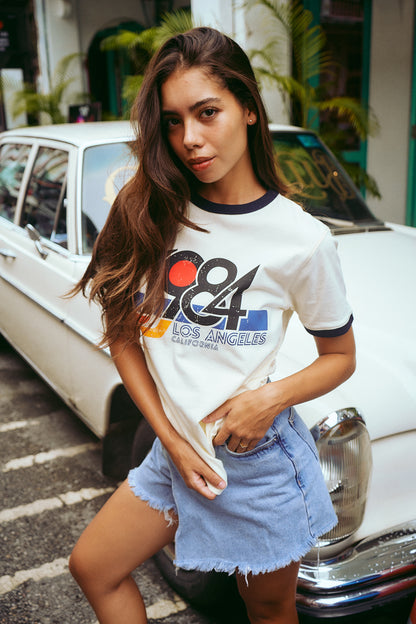 A woman wearing a white ringer T-shirt with a colorful '84 Los Angeles' graphic, paired with denim shorts, standing in front of a vintage white car.