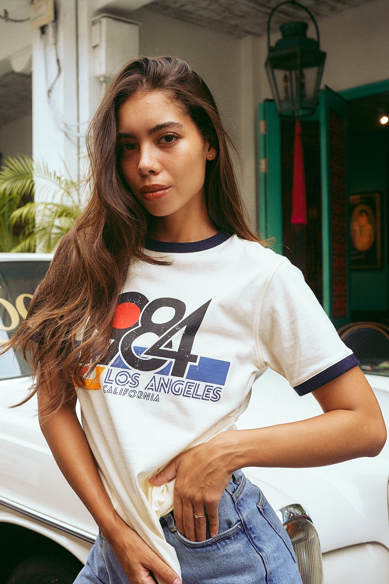 A woman wearing a white graphic t-shirt with navy blue trim and a colorful '68 Los Angeles California' print, paired with light blue jeans, posing outdoors in front of a white vintage car.