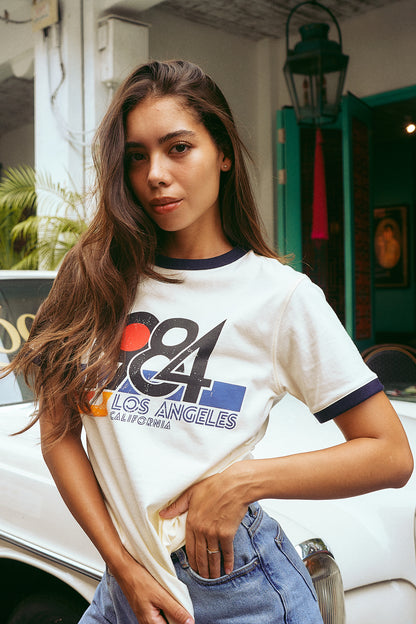 A woman wearing a white graphic t-shirt with navy blue trim and a colorful '68 Los Angeles California' print, paired with light blue jeans, posing outdoors in front of a white vintage car.