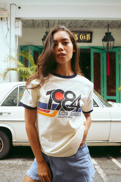 A woman wearing a cream-colored ringer t-shirt with navy blue trim and a colorful graphic print that reads '1984 LOS ANGELES CALIFORNIA'. She is posing outdoors in front of a white vintage car and a building with teal doors.