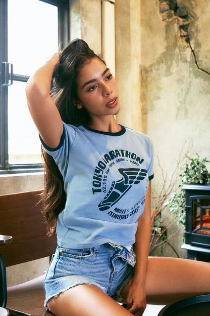 A woman wearing a light blue ringer T-shirt with a dark navy graphic that reads 'TOKYO RANATHON' and 'MUTCH CHADSKIN', paired with denim shorts, sitting in a rustic indoor setting.